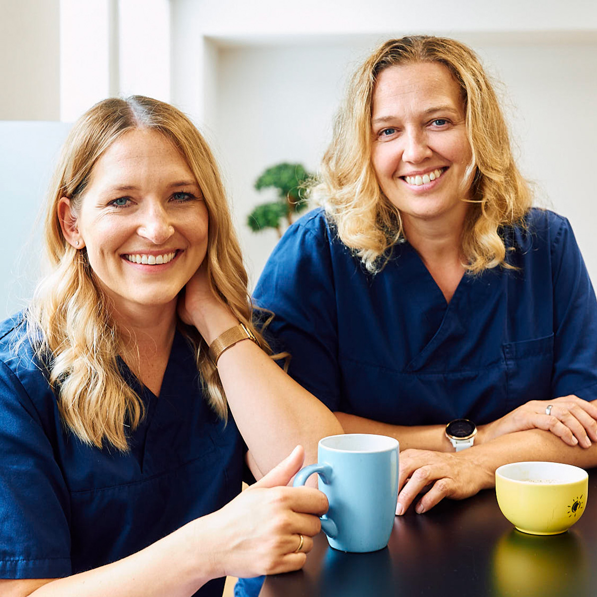 Zwei Zahnärztinnen mit Kaffeetassen lächeln in die Kamera in der Zahnarztpraxis Pfau in Rottweil