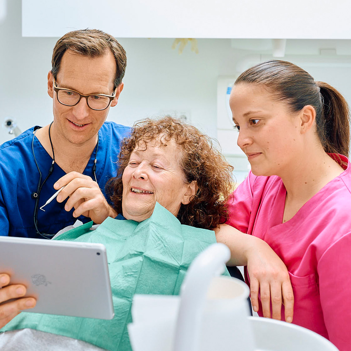 Lachenden Patientin auf dem Zahnarztstuhl, daneben zeigt Zahnarzt Christian Pfau ihr ein tablet, eine ZFA sieht zu in der Zahnarztpraxis Pfau in Rottweil
