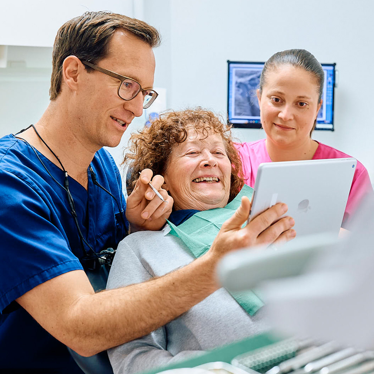 Lachenden Patientin auf dem Zahnarztstuhl, daneben zeigt Zahnarzt Christian Pfau ihr ein tablet, eine ZFA sieht zu in der Zahnarztpraxis Pfau in Rottweil