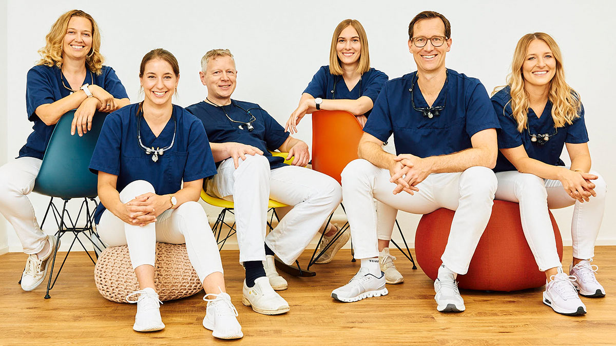 Gruppenbild der Zahnärzte in Rottweil, 6 Personen sitzen lächelnd und entspannt auf Stühlen und Sitzkissen in blauen Shirts und weißen Hosen